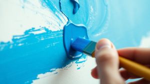 Hand holding paintbrush applying blue paint to canvas.